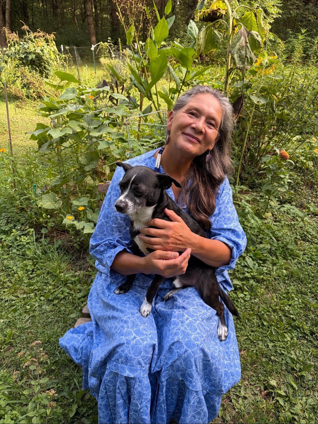 Smiling woman in a blue patterned dress holds a black-and-white dog in a lush garden.